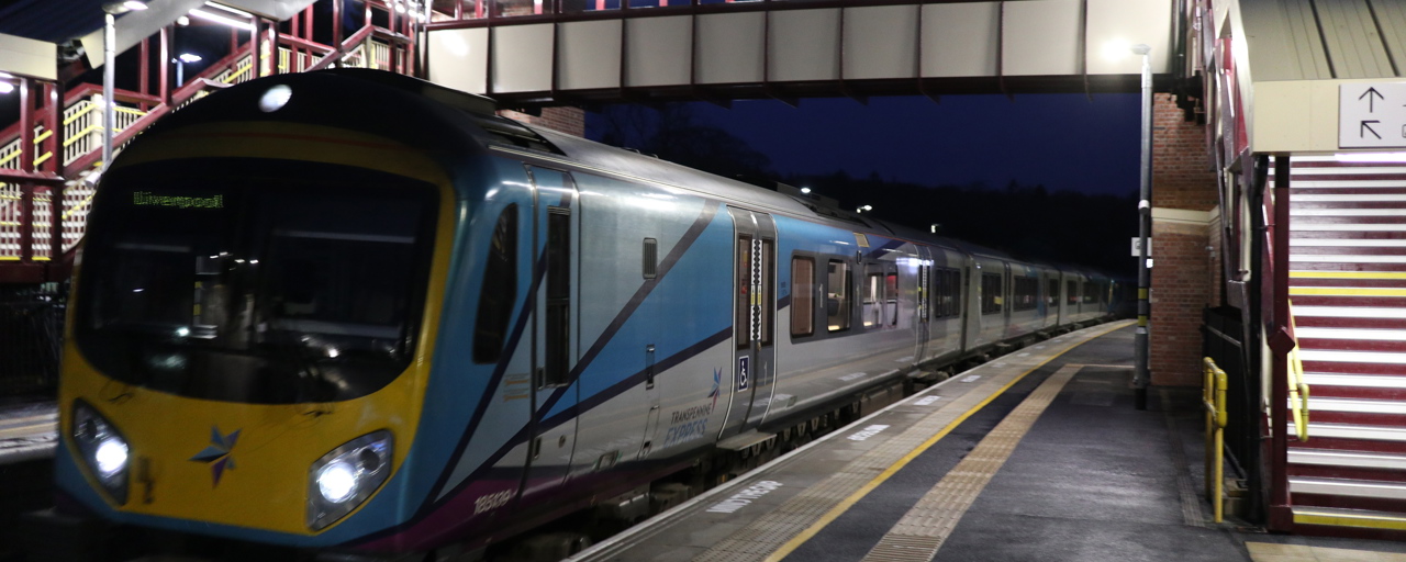 Photo of a passenger train passing under a pedestrian bridge that spans across platforms at a train station