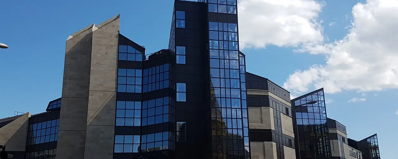 modern building with a distinctive architectural style. The building features a mix of concrete and dark glass facades, with several angular, geometric shapes and pointed rooflines. The structure appears to have multiple sections, including a central tower with a glass facade that reflects the sky and surrounding structures. The sky is clear with a few scattered clouds, providing a bright contrast to the dark glass of the building.