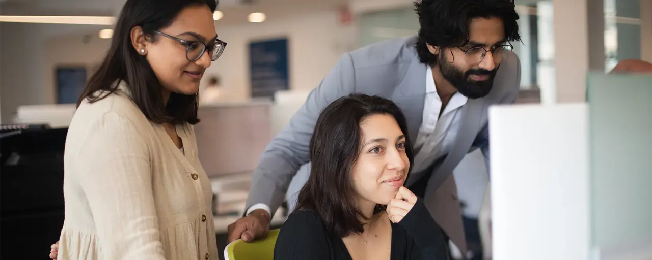 Three colleagues gathered around work desk staring at desktop screen.