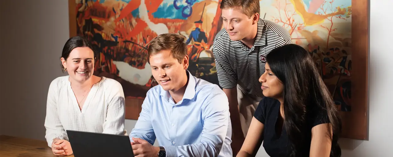 Four colleagues sitting in an office space gathered around male colleague staring at his laptop on a brown table, engaged in a lively conversation. 