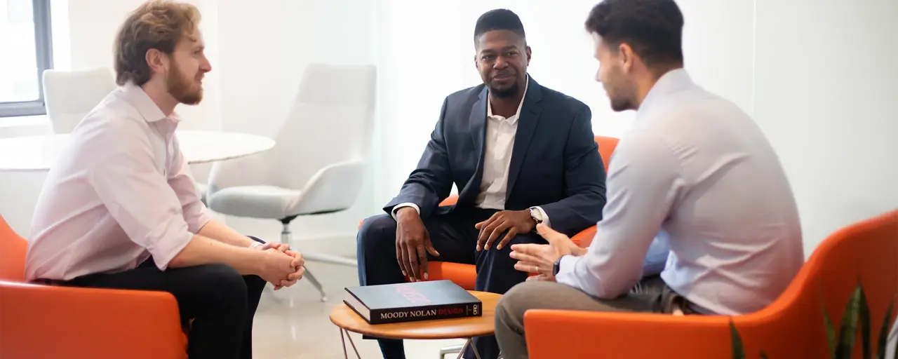 Three male colleagues sitting down on bright orange chairs, engaging in conversation. The office environment is bright and modern with plants behind the chairs.