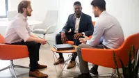 Three male colleagues sitting down on bright orange chairs, engaging in conversation. The office environment is bright and modern with plants behind the chairs.