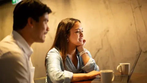 Two colleagues sitting together listening in on conversation with mugs in front of them and yellow wall in the background.