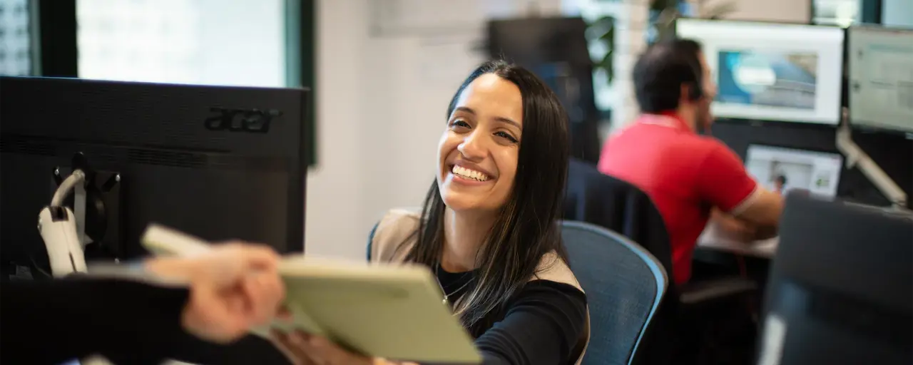 An individual working at a desk handing over a note book to another colleague.