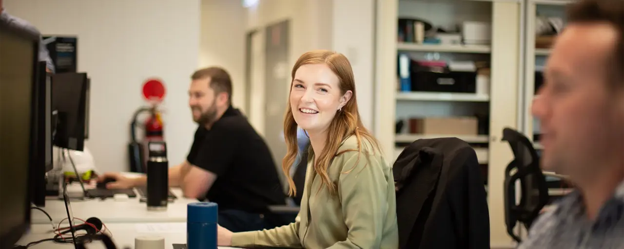  A woman with long hair smiles at the camera while working at her desk. Two male colleagues are working in the background in an open office space with shelves and computers.