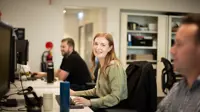  A woman with long hair smiles at the camera while working at her desk. Two male colleagues are working in the background in an open office space with shelves and computers.