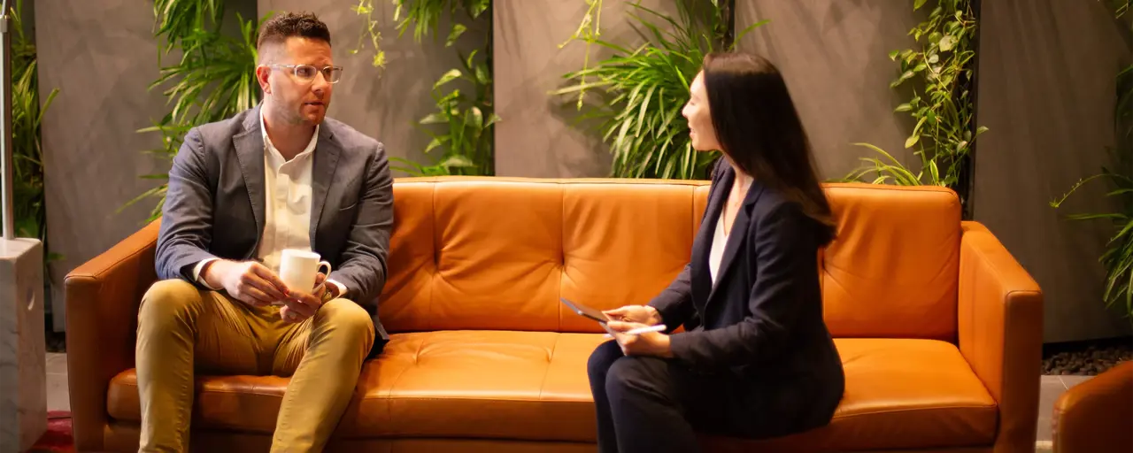 Male and female colleagues sitting together on orange sofas in a plant-filled office having a discussion.