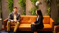 Male and female colleagues sitting together on orange sofas in a plant-filled office having a discussion.