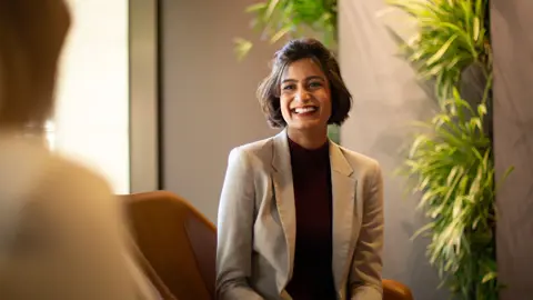 Female employee with short brown hair sitting on orange chair, smiling in a plant-filled office.