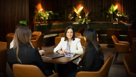 Group of colleagues gathered around brown table talking in a plant-filled office.