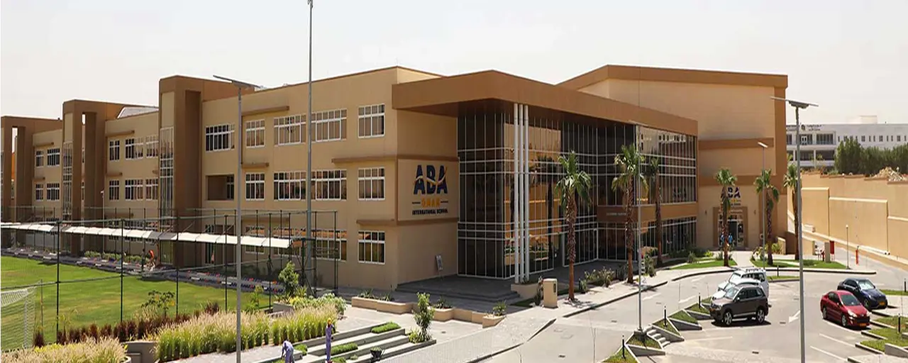 A sand coloured block of school buildings with a car park in front and small playing field to the left of the building. 