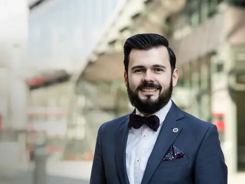 Adrian Predoi, Director, Head of Programme Advisory, Riyadh, Saudi Arabia, in a suit and bow tie smiling outdoors in front of a glass building