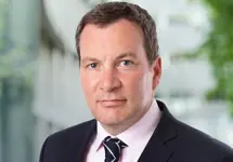 Andy Scott, Managing Director Defence, UK, standing outside in front of an outdoor urban setting, featuring modern architectural elements with clean lines. There are hints of greenery, suggesting some trees or plants.