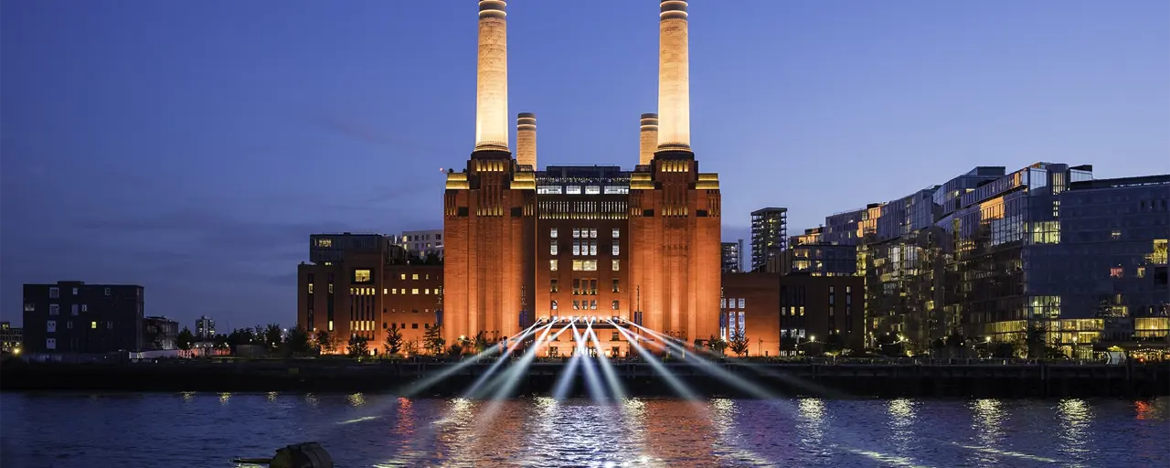 A striking image of London's iconic Battersea Power Station against a blue night sky. The industrial columns are lit up and light also shines on the water in front. To the right of the building are a block of modern, glass fronted flats. 