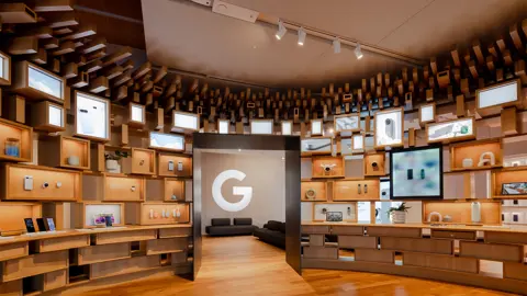 Interior photo of the Google Visitor Experience at Google's Gradient Canopy office building, showing wooden shelves of electronic devices around an archway leading to two dark-grey couches in the background with Google's 'G' logo displayed on a nearby wall