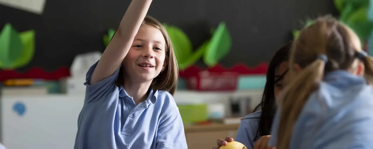 Children together in a classroom with a student smiling whilst putting their hand up