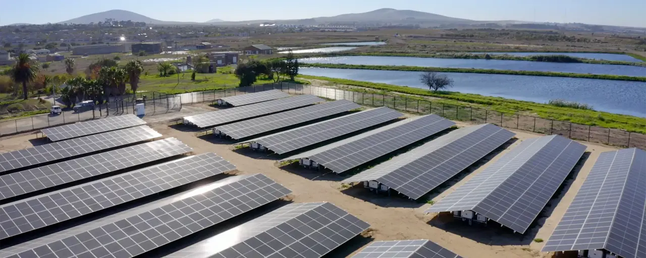 A collection of solar panels next to a body of water with mountains in the background. 