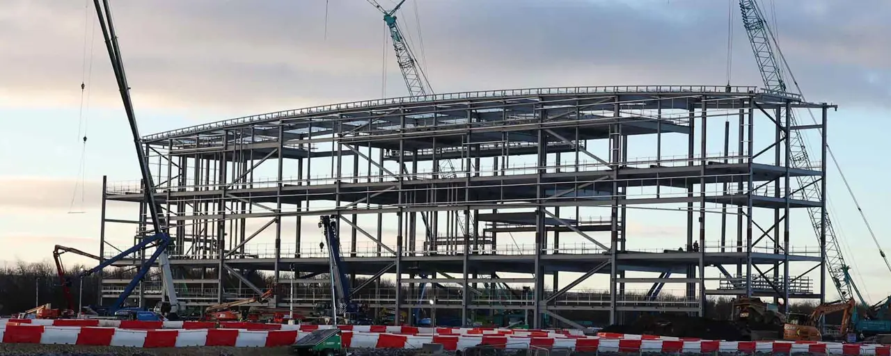 In-progress photo of a factory building to be constructed, with multiple cranes assembling scaffolding