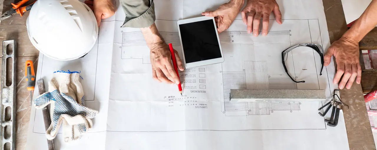 Three work people reading building plans on a table, includes iPad and construction work hat and safety glasses