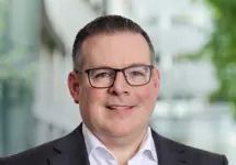  Portrait of Euan Reaper, Director, Europe, Cost Management, standing outside in front of an outdoor urban setting, featuring modern architectural elements with clean lines. There are hints of greenery, suggesting some trees or plants.