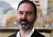 Gonzalo Rosa, Director, standing outside featuring an urban cityscape with tall buildings and a mix of architectural styles. The scene includes some trees with autumn foliage, adding a touch of color. 