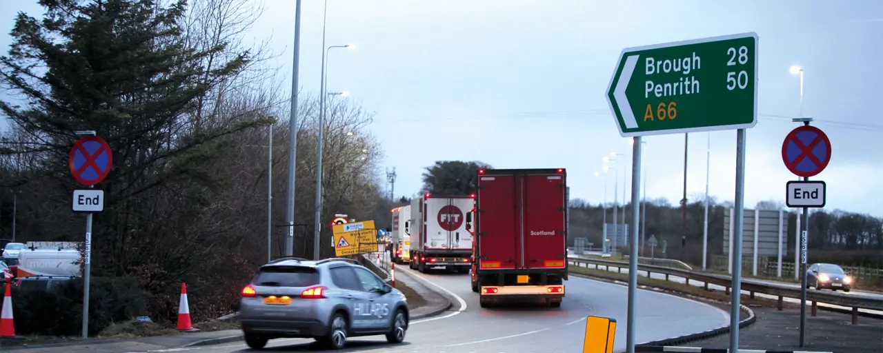 Photo of a car and two trucks joining an offramp on a road with sign on the right of the image indicating A66 route