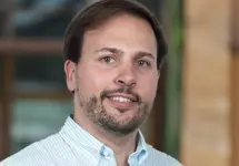 Portrait of Guillermo Martinez, Director, Industrial, Science and Technology, outside in front of a blurred brown background featuring a building and mix of natural and urban elements.