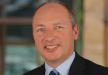  James Hamilton, Director, Global Host Cities and Sports Venues, outside in front of a blurred brown background featuring a building and mix of natural and urban elements.