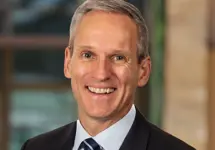 John Robbins, Managing Director, USA and North America Head of Real Estate, outside in front of a blurred brown background featuring a building and mix of natural and urban elements.