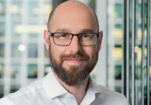 Portrait of Kristoffer Hudson, Associate Director, with city landscape featuring building with lots of windows in the background.