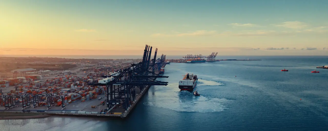 An aerial view of a large shipping port at sunset. The port is lined with numerous container cranes and stacks of shipping containers. A cargo ship is seen navigating out of the port towards the open sea, leaving a wake behind. The sky is clear with a gradient from light yellow to blue as the sun sets in the distance.