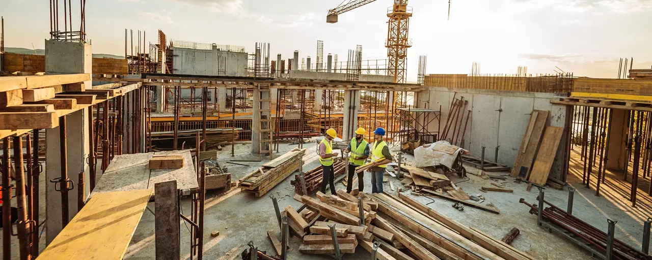 Three male construction workers discussing plans on the floor of a building under construction with a crane in background skyline.