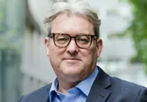 Murray Rowden, Global Head of Infrastructure, standing outside in front of an outdoor urban setting, featuring modern architectural elements with clean lines. There are hints of greenery, suggesting some trees or plants.