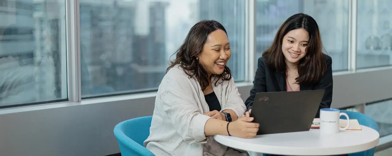 Two female colleagues gathered around table sitting on blue chairs looking at laptop.