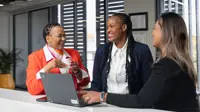 Three female colleagues in office space gathered around laptop, smiling and engaging in conversation.