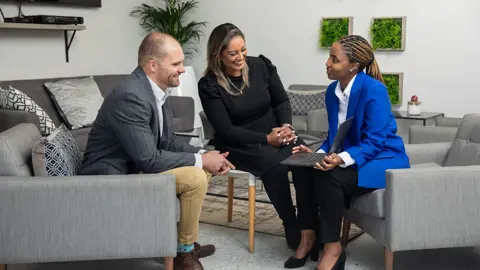 Three colleagues in office sitting down on grey chairs, smiling and engaging in conversation.