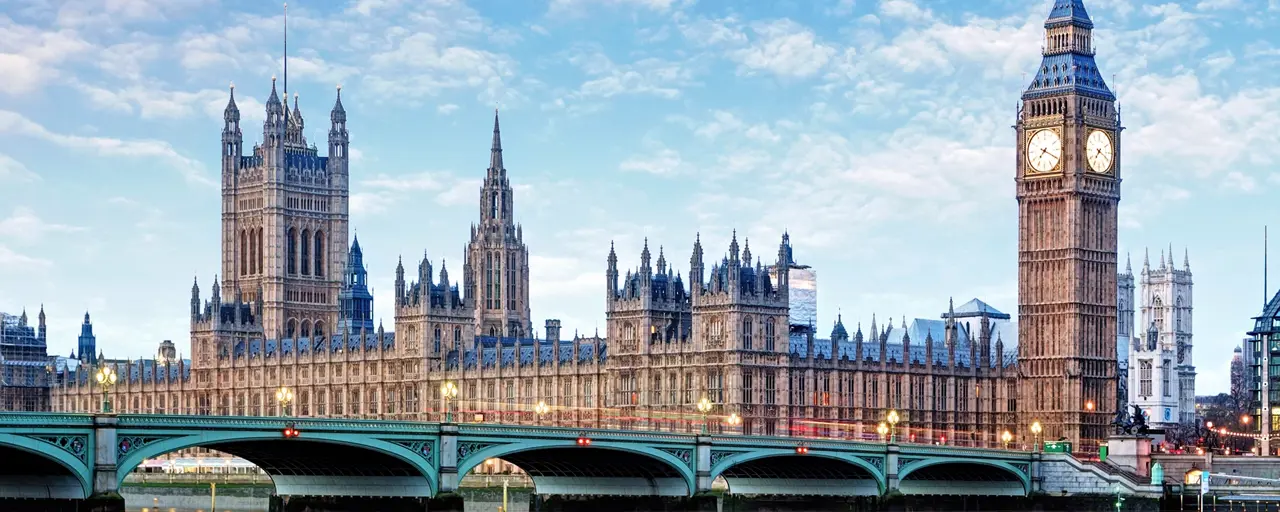 Houses of Parliament, in London, England. Taken from across the River Thames, capturing the entirety of the historic building, including the Elizabeth Tower, which houses Big Ben, on the right. The Westminster Bridge spans the river in the foreground, with the calm water reflecting the structure above. The sky is partly cloudy, and the scene is bathed in soft daylight.