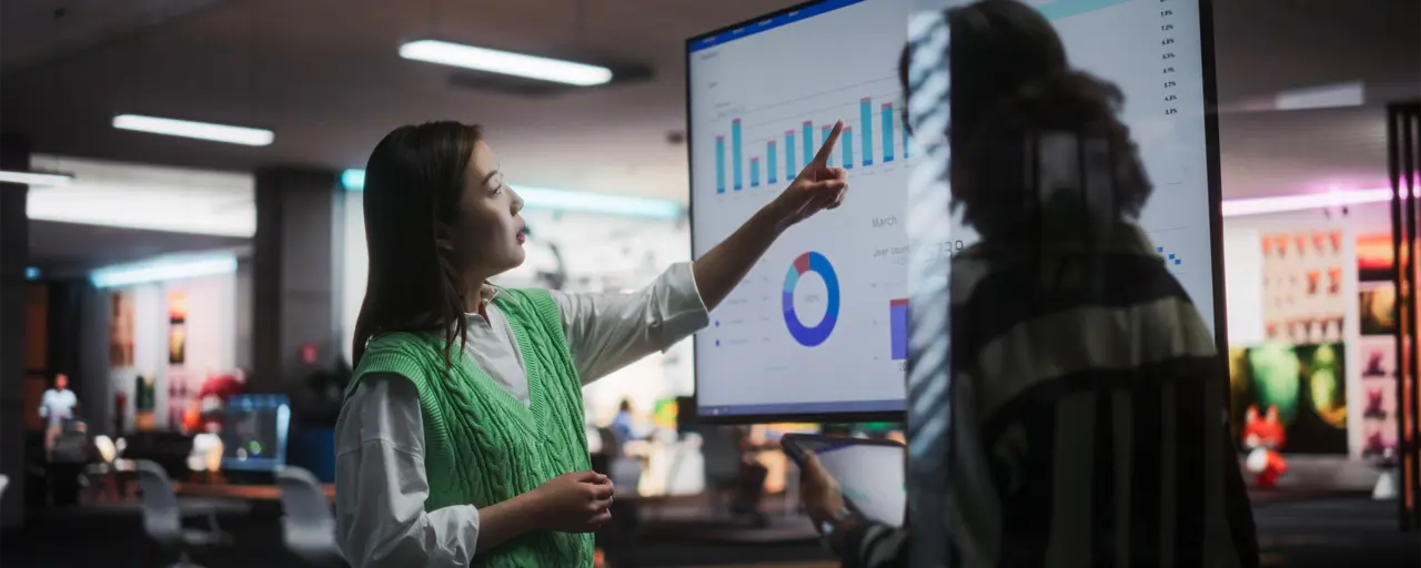 Two women in an office pointing at a screen with charts on it