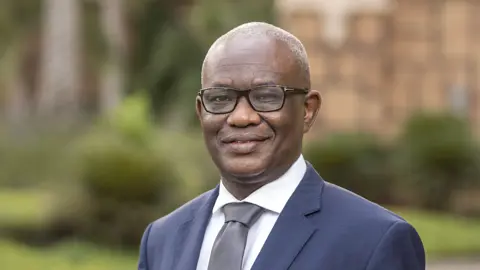 Portrait of Segun Faniran, Forensic Planning and Delay, Middle East, outside in front of grass and bushes with tall buildings behind.