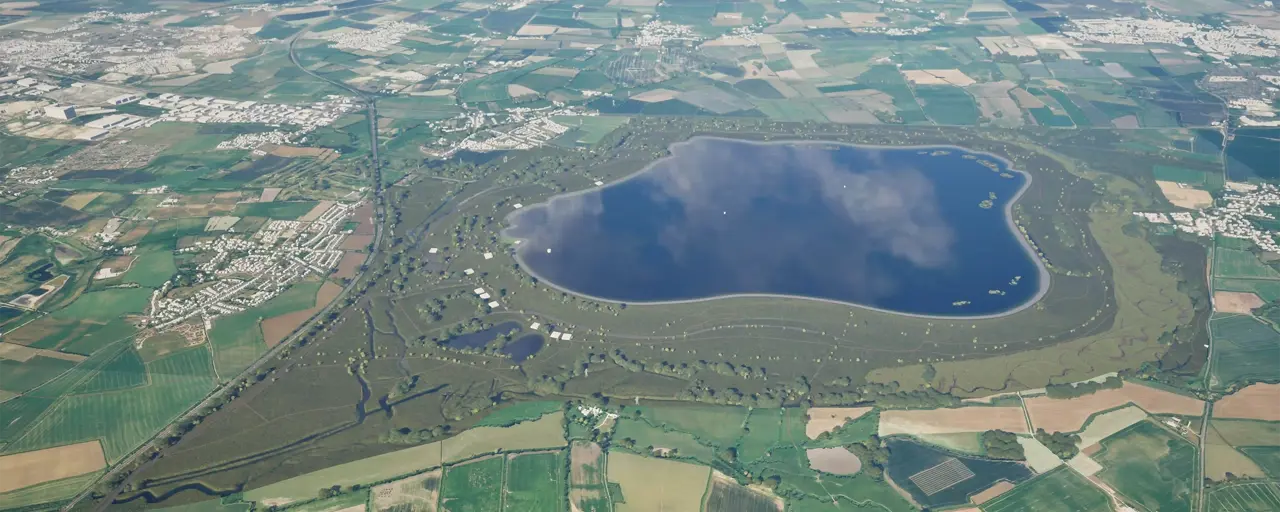 An aerial view of a large stone dam with a reservoir on the right side and a lush green landscape surrounding it. A small building is visible near the base of the dam, adjacent to a body of water and rocky terrain.