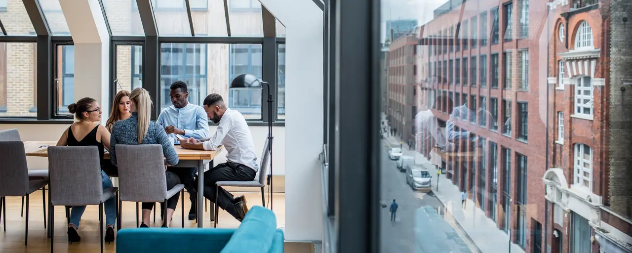 A group of male and female work colleagues sitting at a boardroom desk with exterior street view to the left