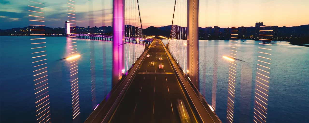 View from a bridge suspended over water at dusk with city lights in the distance