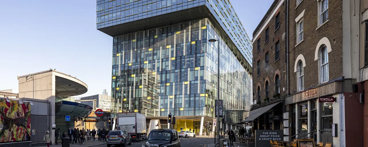 Modern urban street scene in London, featuring a striking contemporary glass building with a cantilevered upper section. This building is set against a backdrop of older brick structures, including a corner restaurant named "Tortilla" with outdoor seating. A classic black London taxi and other vehicles are visible on the street, along with pedestrians walking by. The area appears busy with a mix of architectural styles, reflecting both modern and historical elements of the city. 