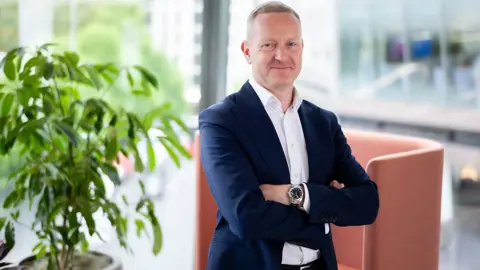 Portrait of Neil Bullen, Global Managing Director, Real Estate, In office standing by plant with his arms crossed.