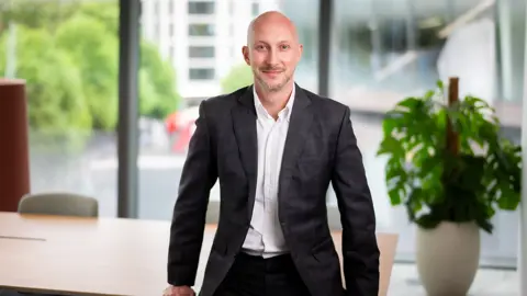 Portrait of James Hardy, Director, Head of UK Sustainability, standing in office space next to plant.