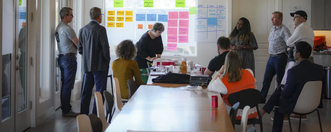 Photo of a group of people seated and standing around a table in a boardroom during a meeting, with a project screen behind them displaying different coloured blocks of text in the form of an organisational chart