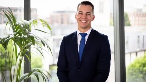 Portrait of Nicolas Offroy, Principal Consultant, Australia, standing in office space against window and plants.