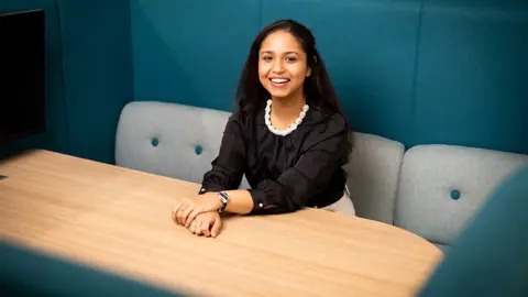 Portrait of Christina Joby Maria, Junior Consultant, Sustainability, United Kingdom, sitting in office space, smiling.