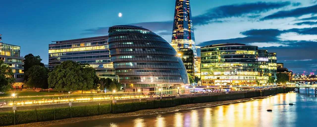 Nighttime skyline view of the South bank of the Thames River in London, with the City Hall building in the foreground