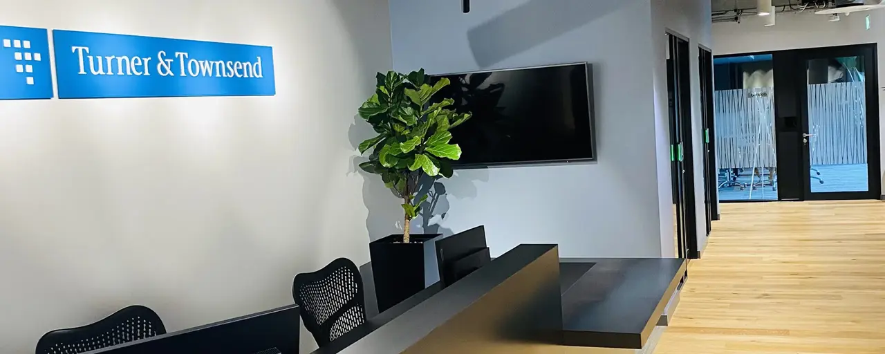 Picture of office reception and hallway with the sign 'Turner & Townsend' above reception chairs. There is a plant in the corner in front of a tv.